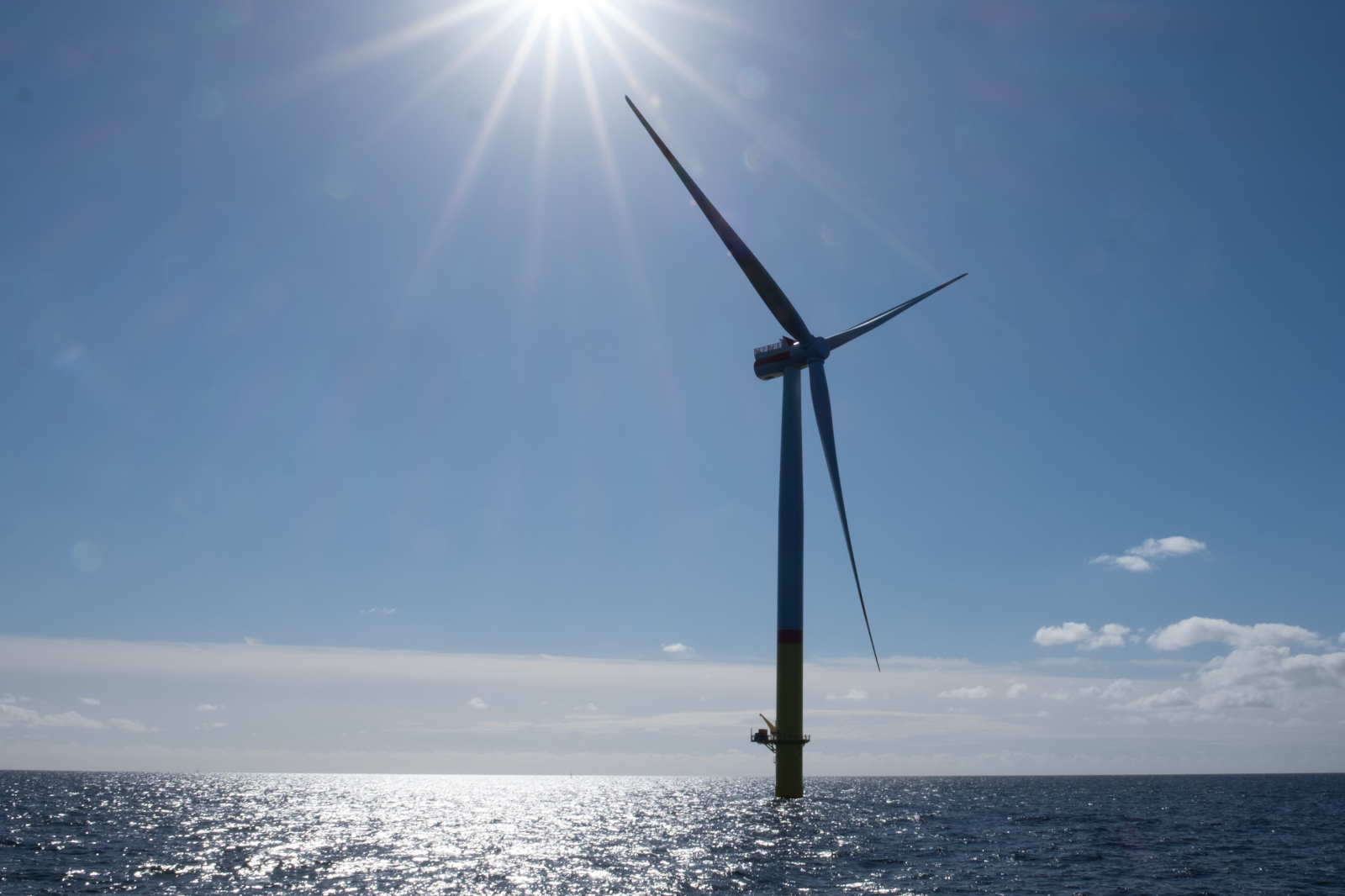 A towering offshore wind turbine stands against a bright blue sky, with sunlight glinting off the ocean waves below.