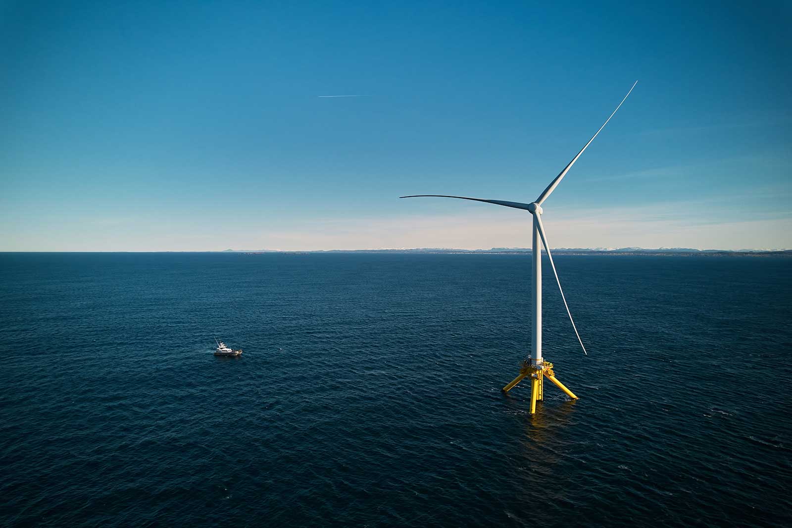 A large wind turbine stands in the ocean with a boat nearby and mountains visible in the distance under a clear blue sky.